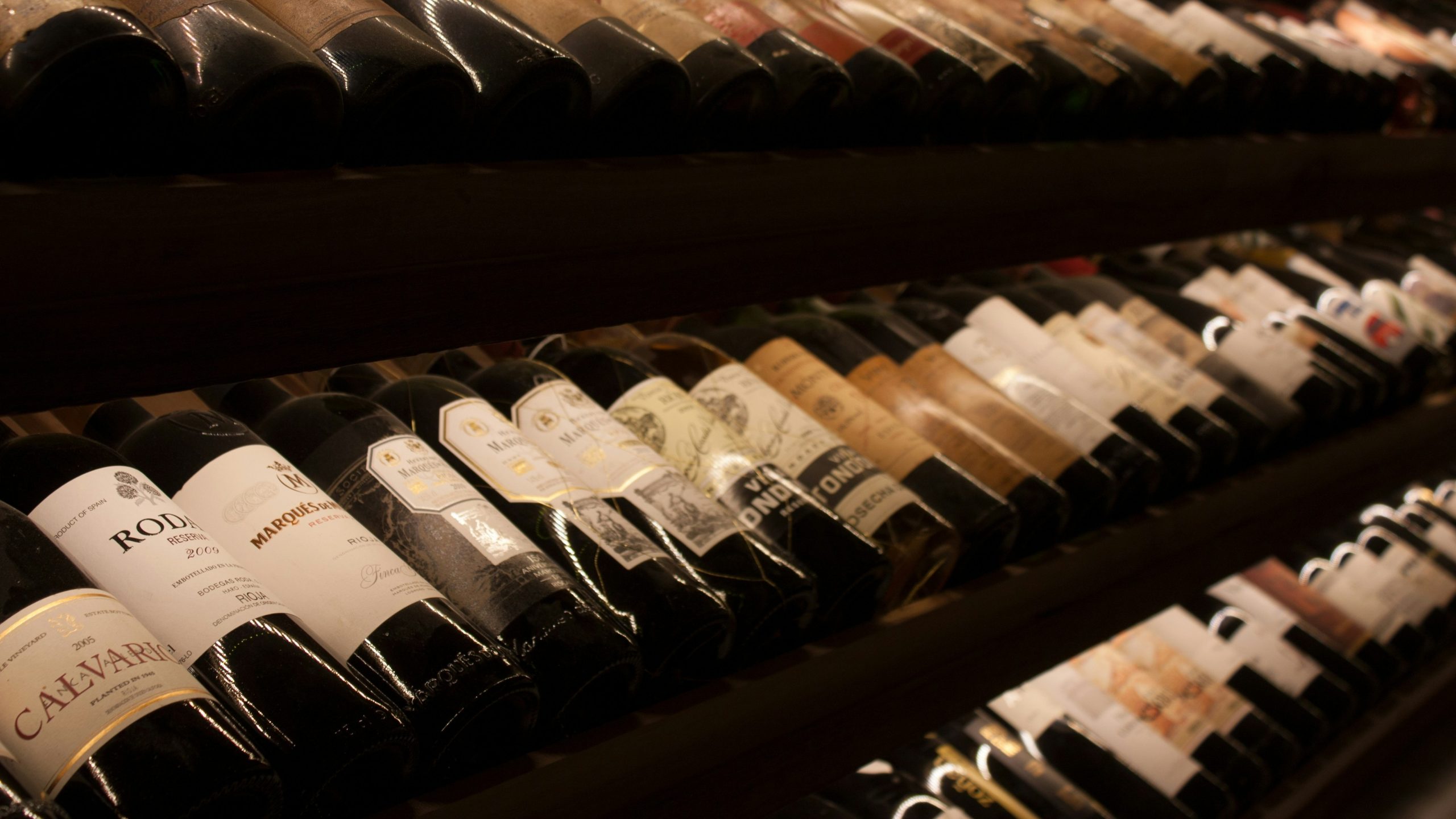 File di bottiglie di vino assortite appoggiate sui fianchi su scaffali di legno in una cantina poco illuminata, con etichette di vari colori e coperchi di stagnola visibili.