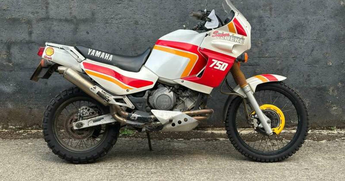 Una moto d'epoca Yamaha XTZ 750 Super Ténéré con grafiche rosse, bianche e gialle è parcheggiata su una superficie di cemento di fronte a un muro grigio.
