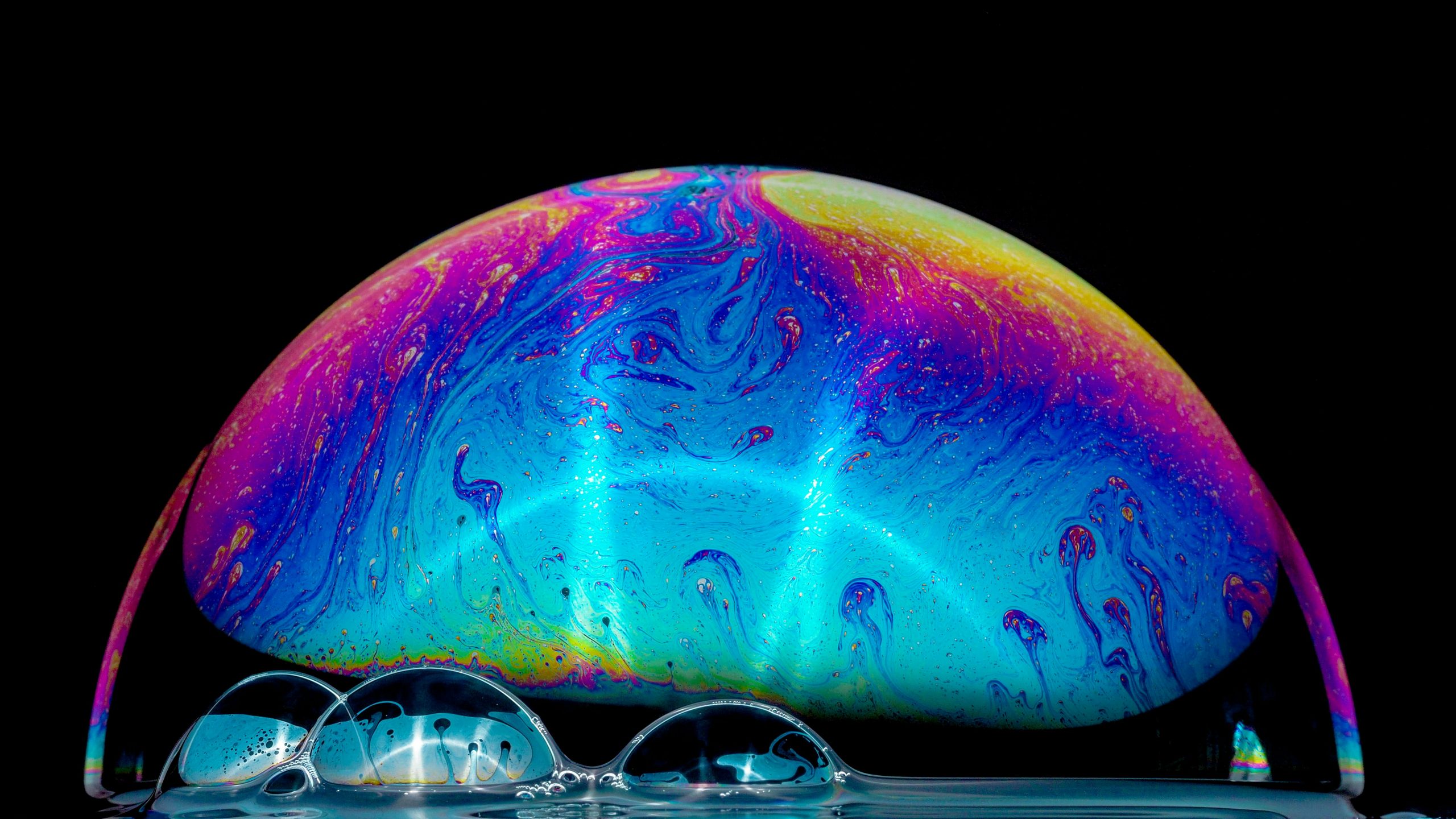 Primo piano di una cupola di bolle di sapone con bolle più piccole al di sotto, che mostra vivaci vortici iridescenti di blu, rosa, arancione e giallo su uno sfondo nero.