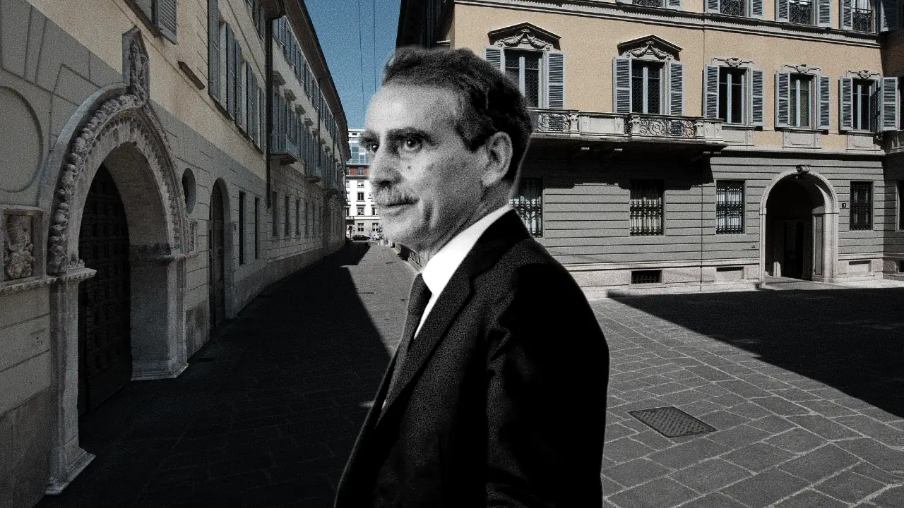 Un uomo in abito scuro si trova in una strada acciottolata fiancheggiata da edifici storici europei e guarda di lato. L'immagine è parzialmente in bianco e nero, in contrasto con lo sfondo colorato.