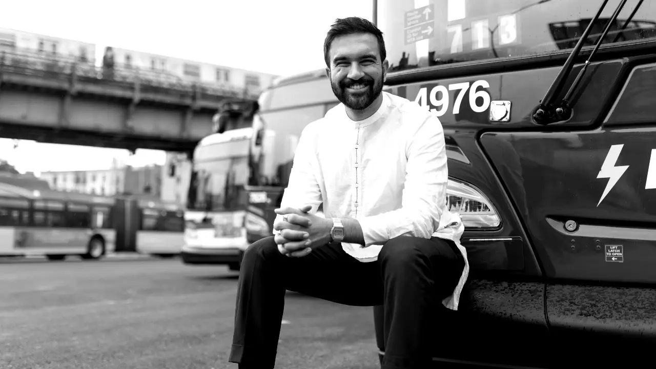 Un uomo sorridente con la barba, che indossa una camicia chiara e pantaloni scuri, siede sul bordo anteriore di un autobus urbano numerato 4976, con altri autobus e un binario ferroviario sopraelevato sullo sfondo.