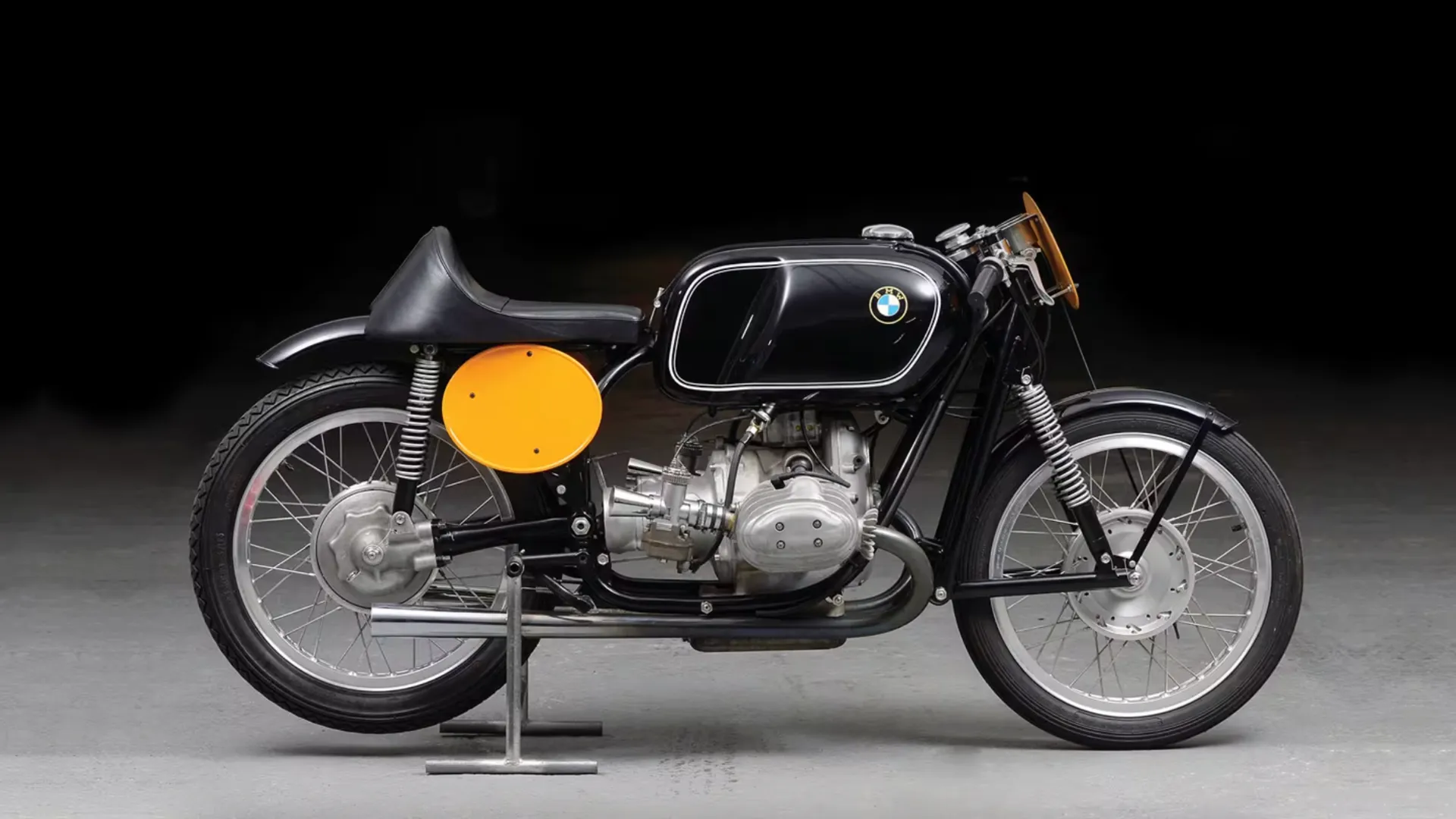 Una moto da corsa BMW nera d'epoca con un grande serbatoio, una targa da corsa gialla e un motore a vista è esposta su un cavalletto su uno sfondo scuro.