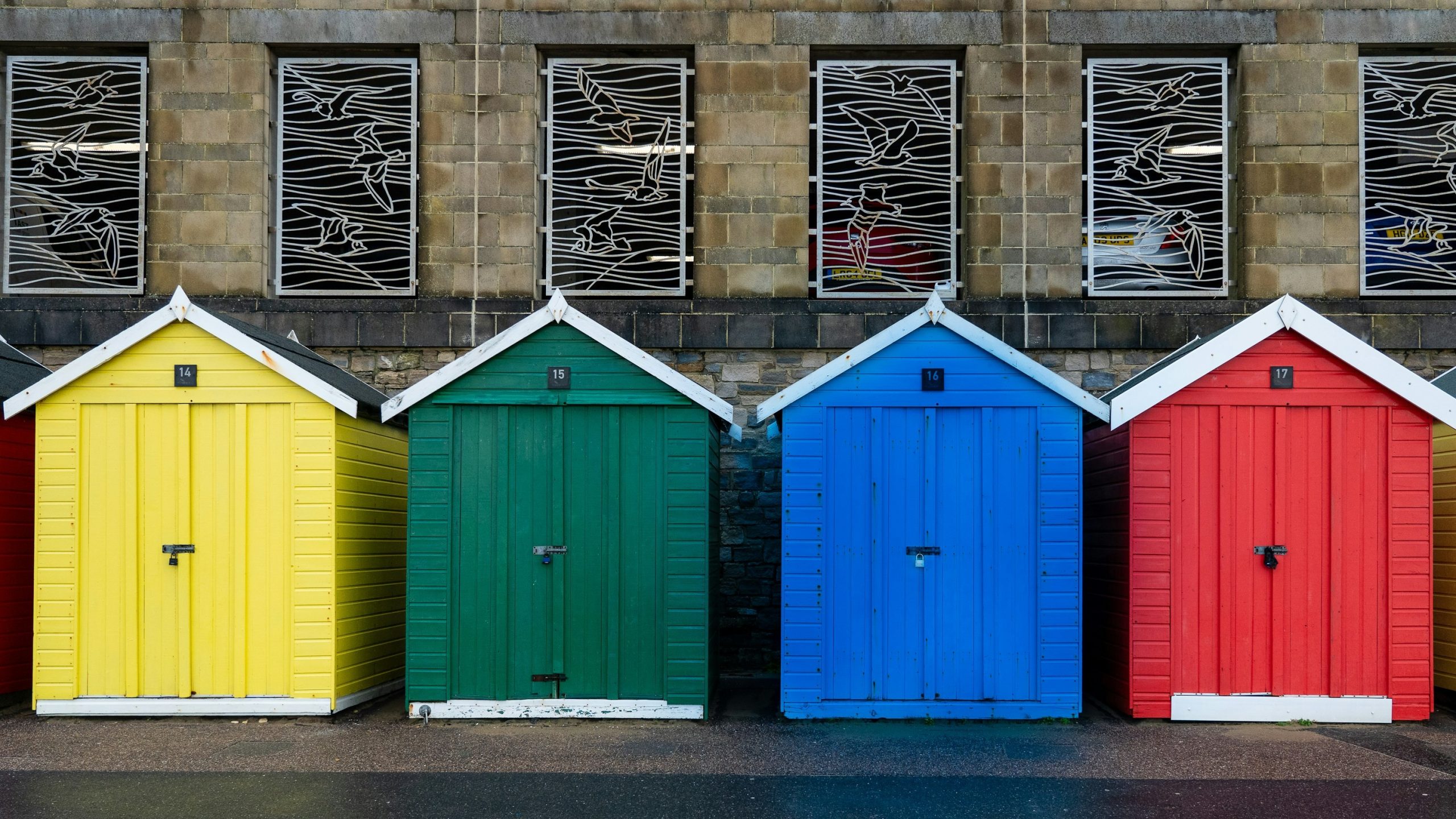 Quattro capannoni di legno colorato - giallo, verde, blu e rosso - si trovano uno accanto all'altro di fronte a un edificio in mattoni con grate bianche decorative alle finestre. Il terreno sembra bagnato, il che fa pensare a una pioggia recente.