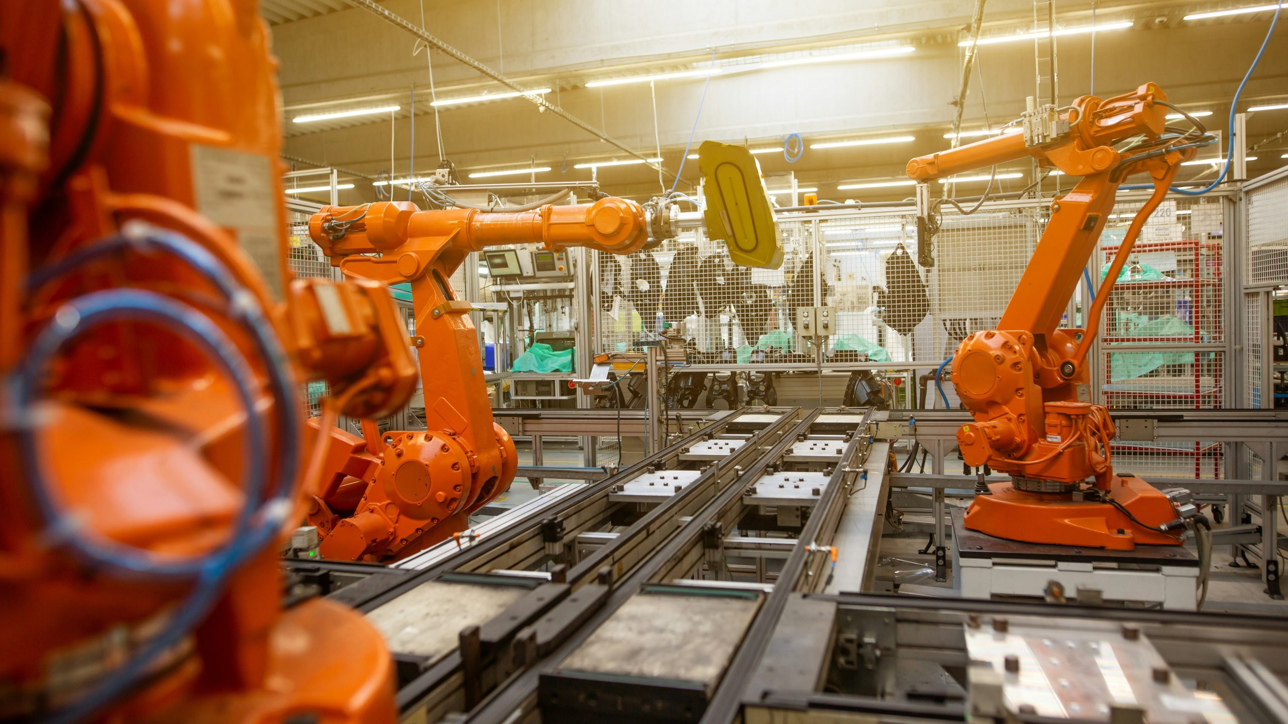 Bracci robotici industriali arancioni lavorano su una linea di assemblaggio automatizzata in una fabbrica moderna, con macchinari e attrezzature visibili sullo sfondo sotto una luminosa illuminazione dall'alto.