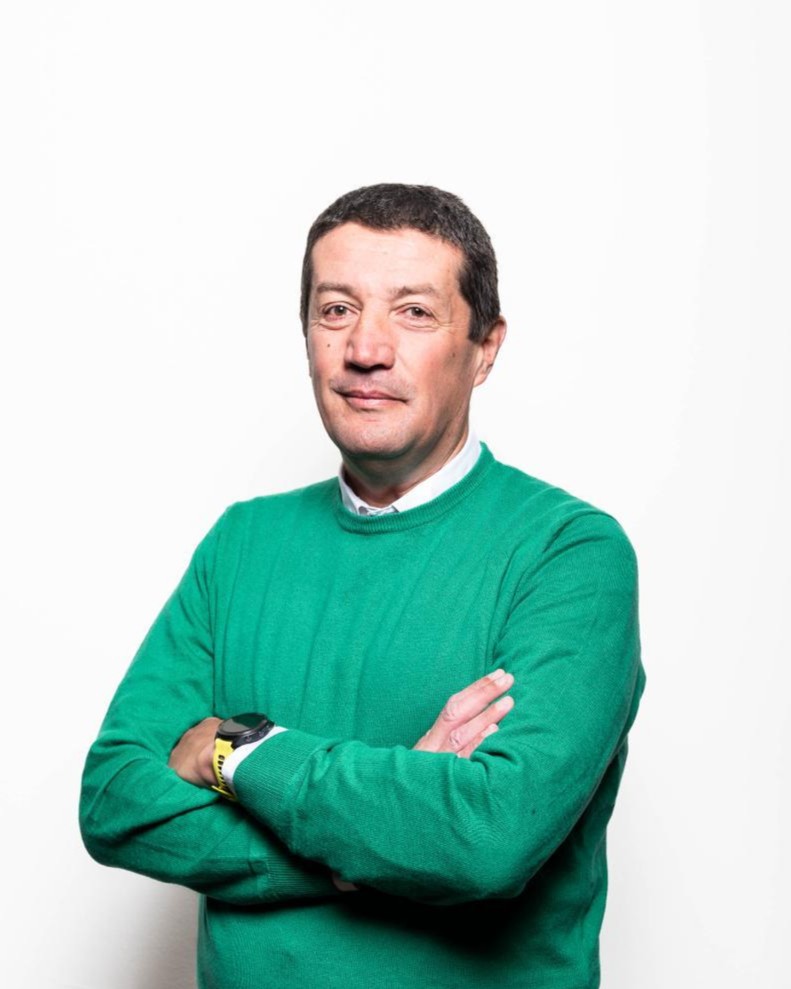 Un uomo di mezza età con i capelli corti e scuri si staglia su uno sfondo bianco, indossando un maglione verde brillante su una camicia bianca. Ha le braccia incrociate e guarda la telecamera con un'espressione neutra.