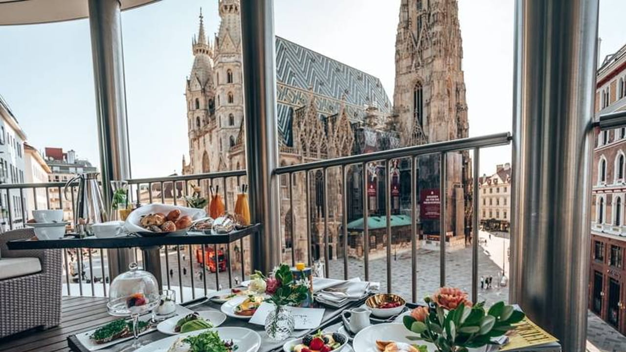 A table set with a variety of breakfast dishes on a balcony overlooking St. Stephen's Cathedral in Vienna, with historic buildings and the cathedral’s ornate facade visible in the background.