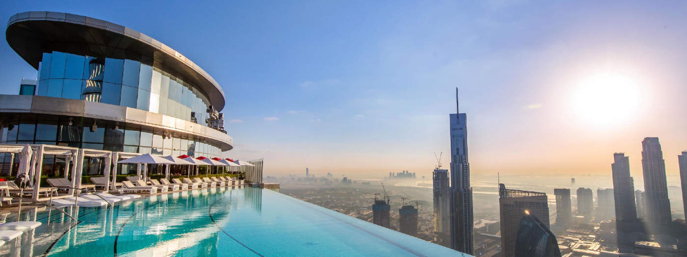 A rooftop infinity pool with lounge chairs overlooks a modern city skyline at sunrise, with tall skyscrapers and a clear sky in the background.