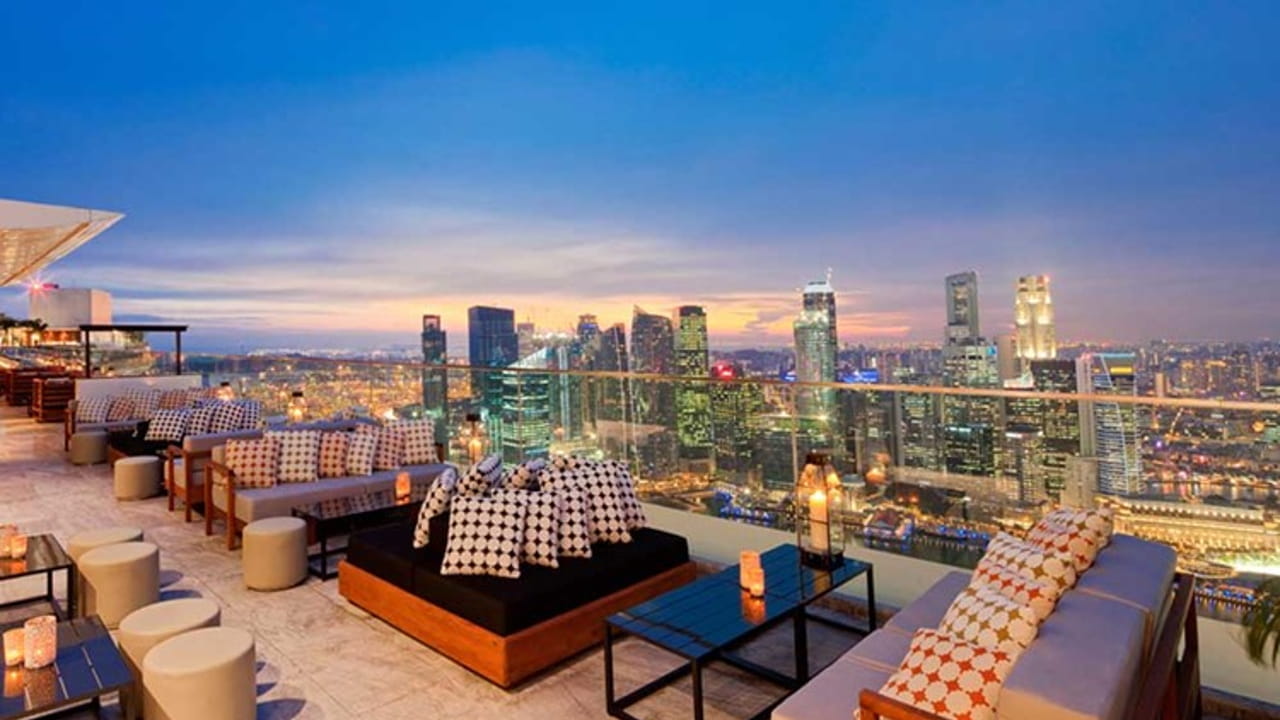 A stylish rooftop bar with modern seating, patterned tablecloths, and glowing lanterns overlooks a city skyline at sunset, with tall buildings and lights illuminating the evening.