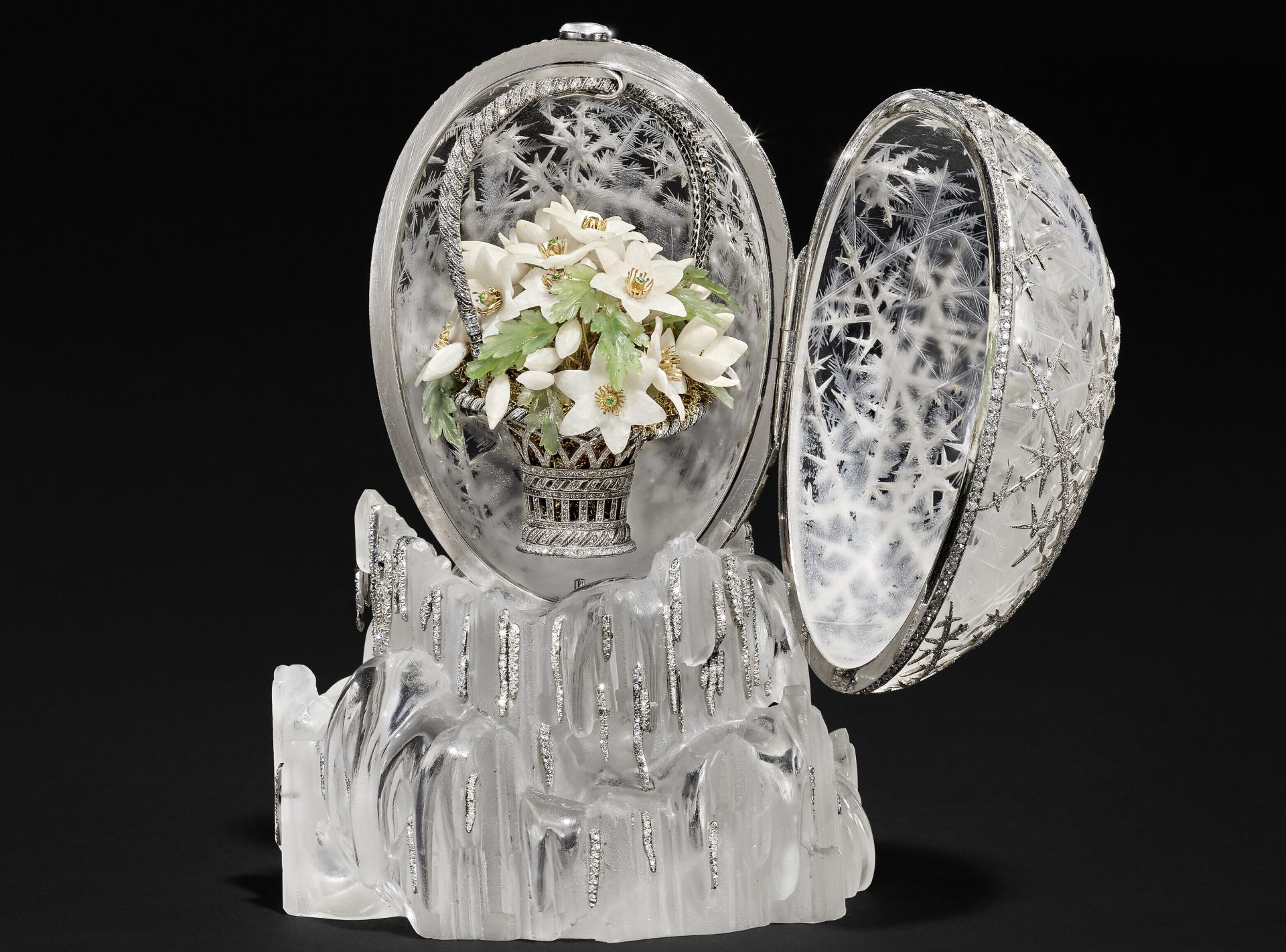 A jeweled egg, decorated with silver snowflake patterns, is open to reveal a basket of white flowers inside. The egg rests on a frosted, ice-like crystal base against a black background.