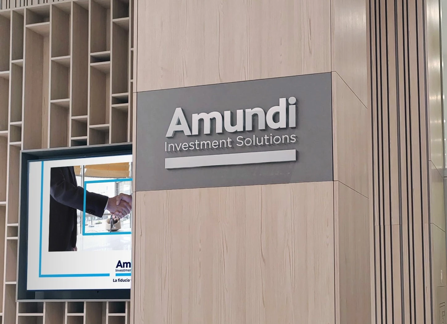 Una parete moderna presenta il logo "Amundi Investment Solutions" in lettere d'argento. Accanto c'è uno schermo che mostra due persone che si stringono la mano con il logo Amundi visibile nell'angolo.