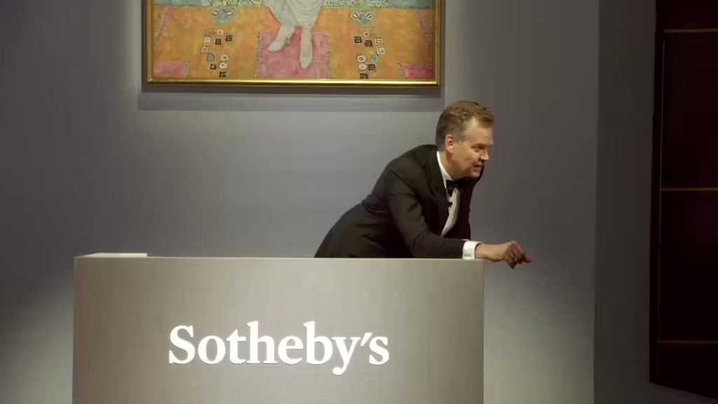 Un banditore di Sotheby's in smoking nero si china sul podio durante un'asta. Un quadro incorniciato con motivi colorati e una figura seduta è appeso alla parete dietro di lui.