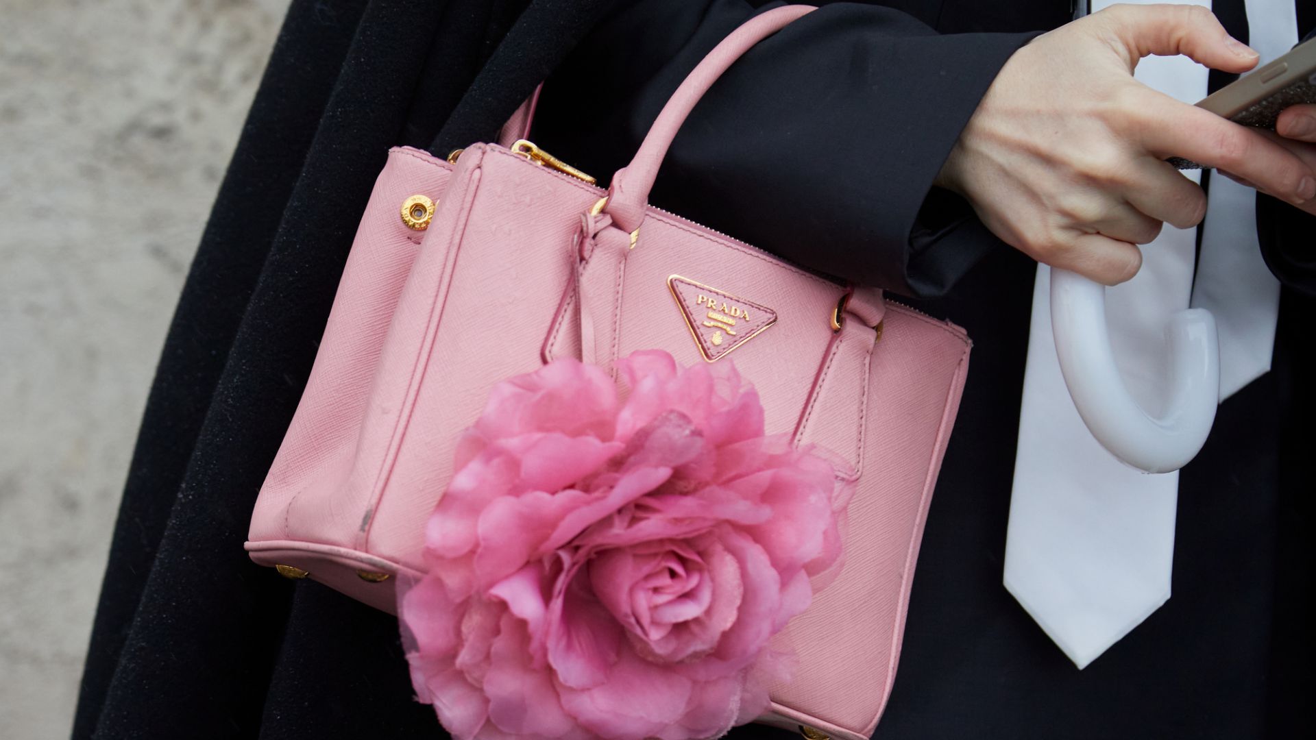 Una persona tiene in mano un telefono e un ombrello mentre trasporta una borsa Prada rosa ornata da un grande fiore rosa. La persona è vestita con un cappotto nero e una cravatta bianca.