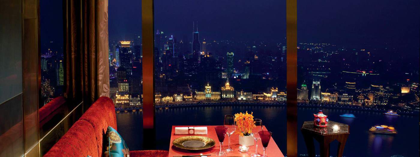 A romantic dinner table for two is set by a window with red chairs, wine glasses, and flowers, overlooking a city skyline and river illuminated by colorful lights at night.