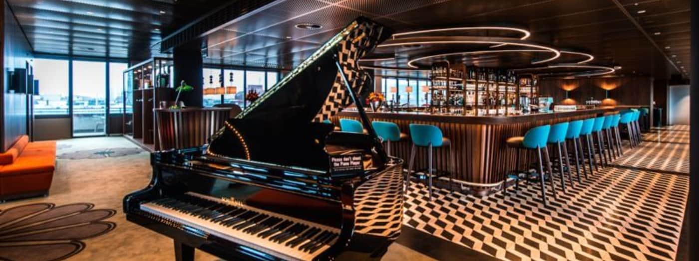A stylish lounge features a glossy grand piano, patterned black-and-white flooring, a modern bar with turquoise barstools, circular lighting, and large windows providing city views.