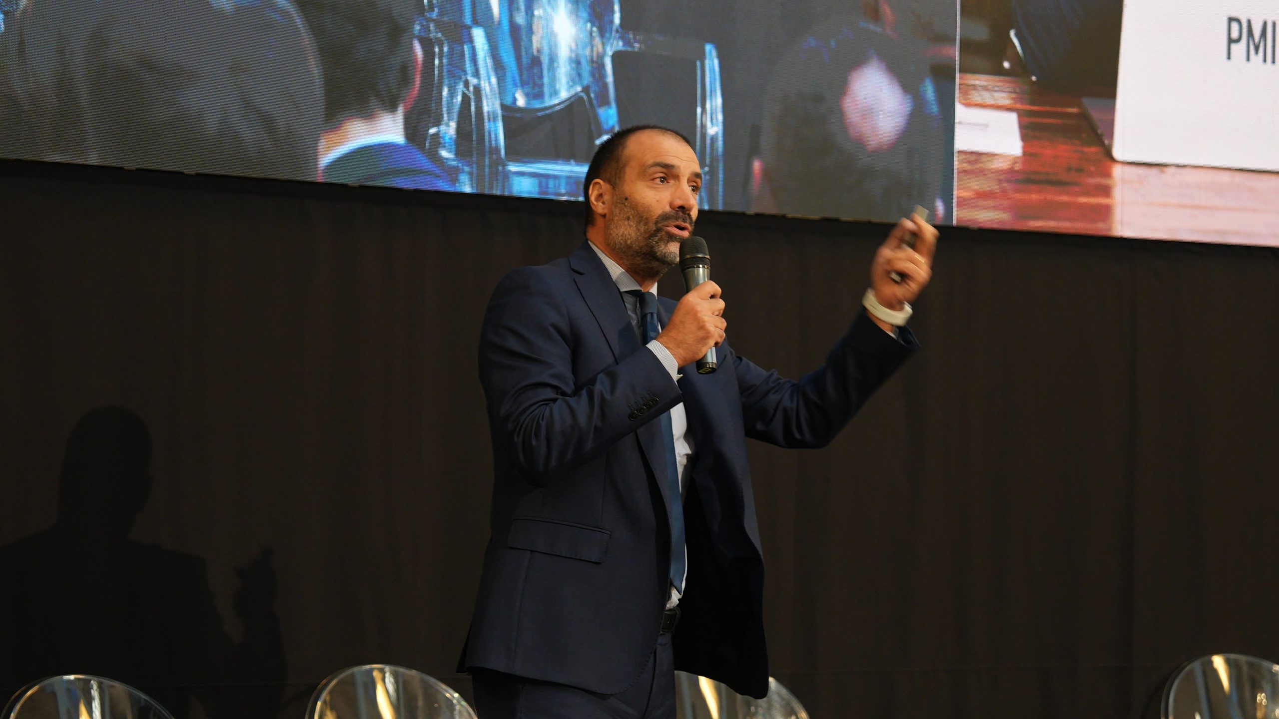Daniele Colantonio di Anthilia, in occasione del Wealth Management Summit, in abito blu tiene un microfono e gesticola mentre parla sul palco del debito privato di Anthilia, davanti a una tenda nera e a un grande schermo che mostra parte del pubblico.