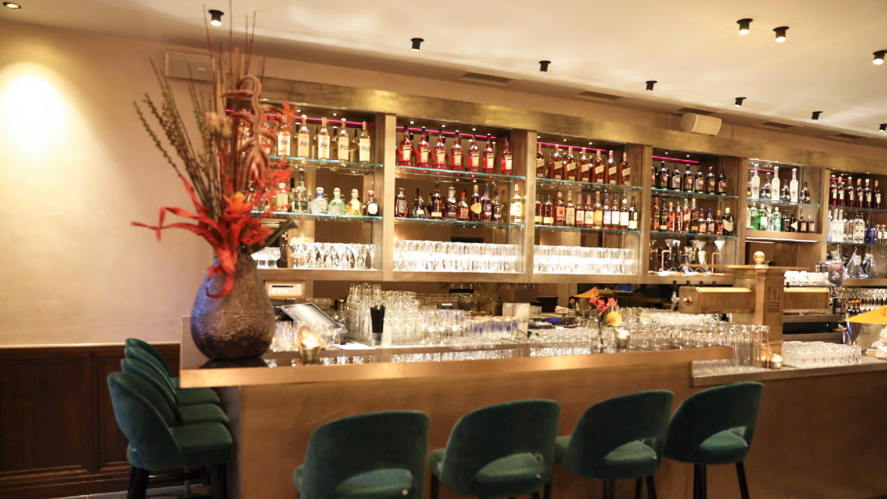 A modern bar with green velvet barstools, a gold counter, glassware, and shelves lined with various liquor bottles. A large decorative vase with red flowers sits on the counter. Warm lighting creates a cozy atmosphere.