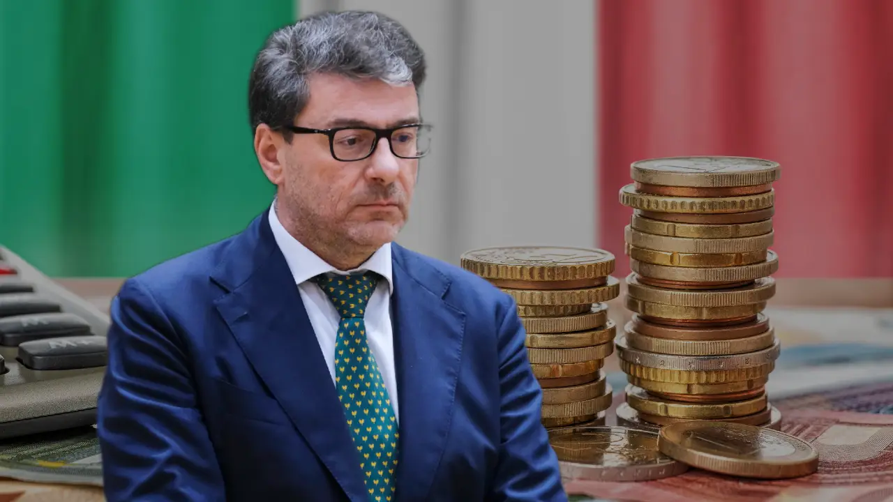 Un uomo in giacca e cravatta e occhiali è seduto davanti a monete impilate e a una calcolatrice, su uno sfondo sfocato con i colori della bandiera italiana.
