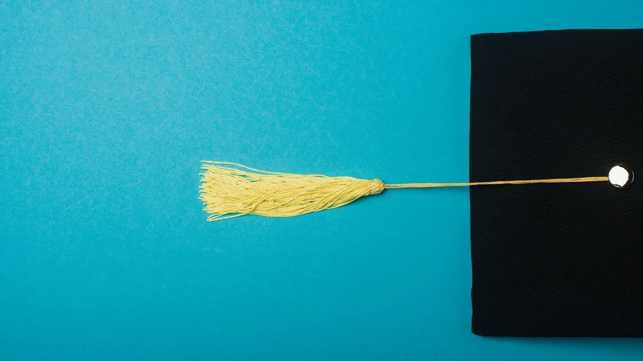 Un cappello di laurea nero con una nappina dorata si trova su uno sfondo blu brillante, con la maggior parte del cappello sul lato destro dell'immagine.
