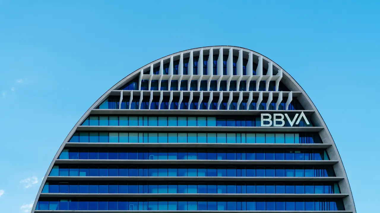 Moderno edificio per uffici con tetto curvo, finestre orizzontali in vetro colorato di blu e la scritta "BBVA" in alto a destra, sullo sfondo di un cielo azzurro.