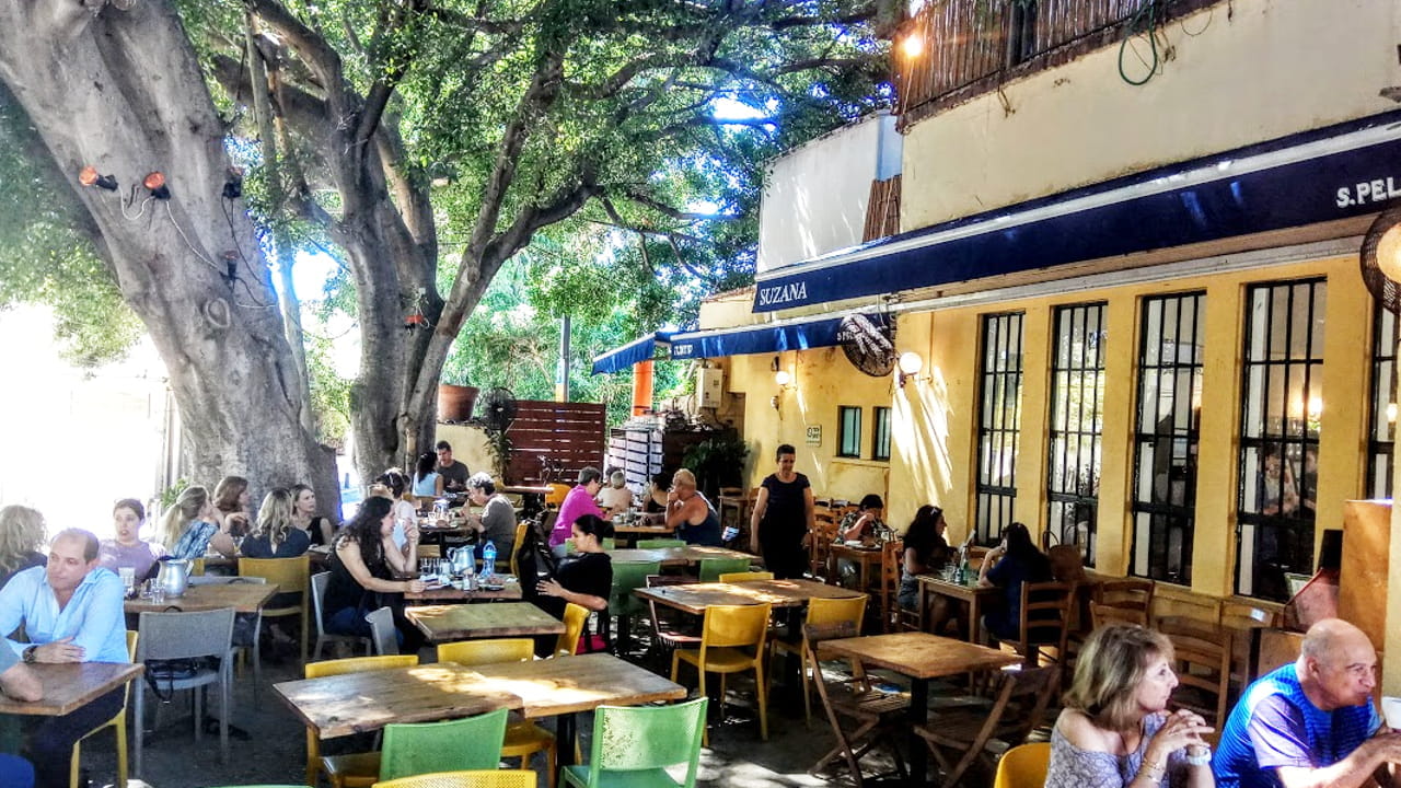 People sit at wooden tables under large shady trees outside a busy café with a blue awning on a sunny day. Some are eating or chatting, while others appear to be waiting or relaxing.