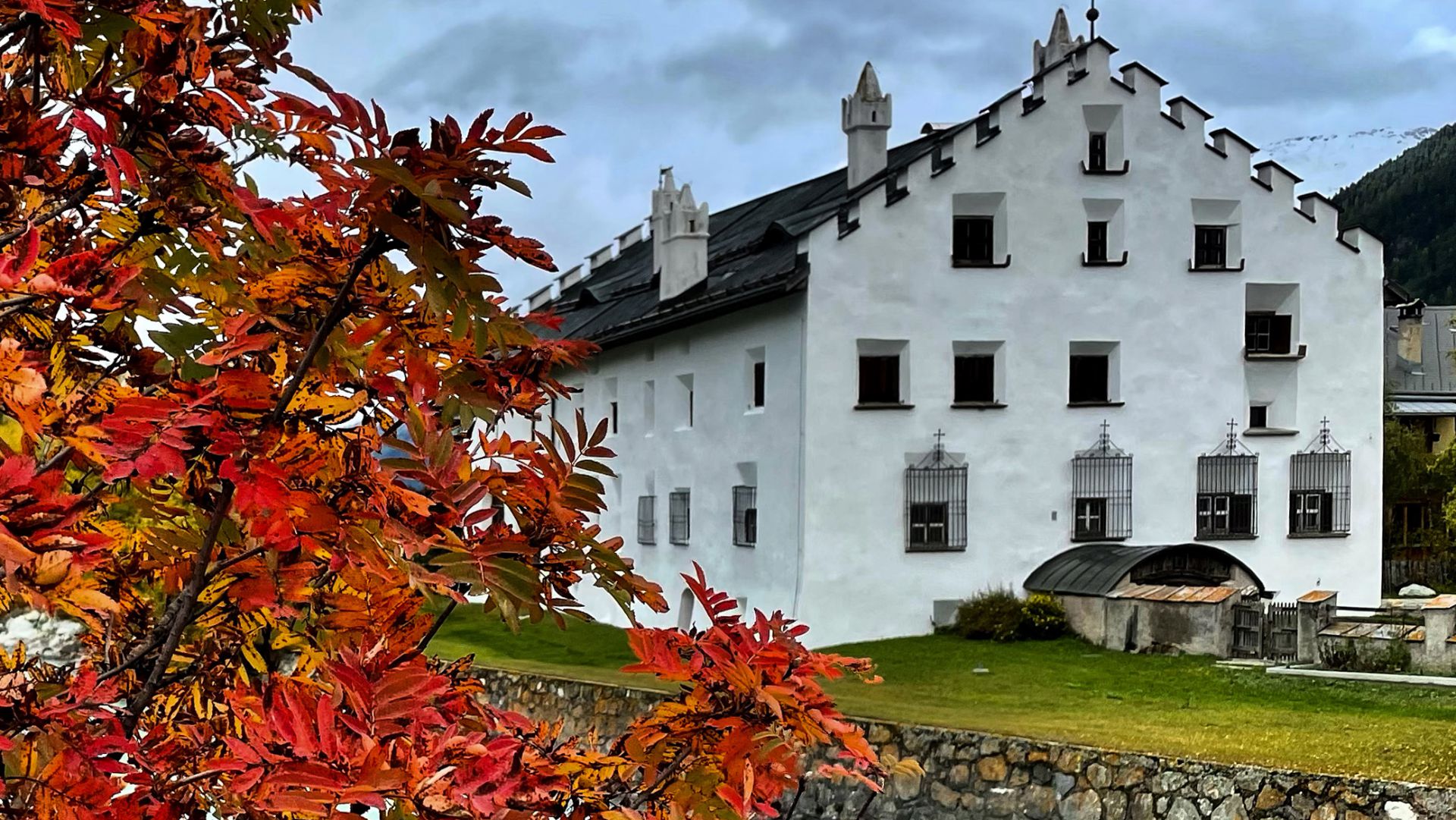 Un edificio storico bianco con frontoni a gradini si erge dietro un muro di pietra, con foglie autunnali di colore rosso e arancione in primo piano sotto un cielo nuvoloso.