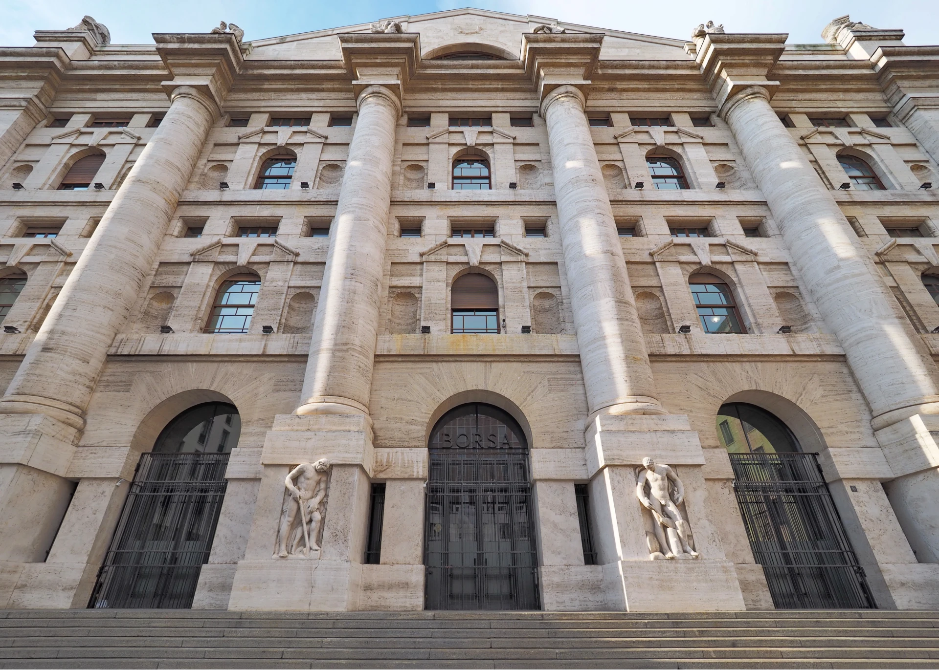 Sotto un cielo terso si staglia la facciata di un imponente edificio in pietra con alte colonne, portali ad arco, sculture vicino agli ingressi e ampi gradini di salita, che evocano il prestigio storico spesso associato all'azionario italiano.