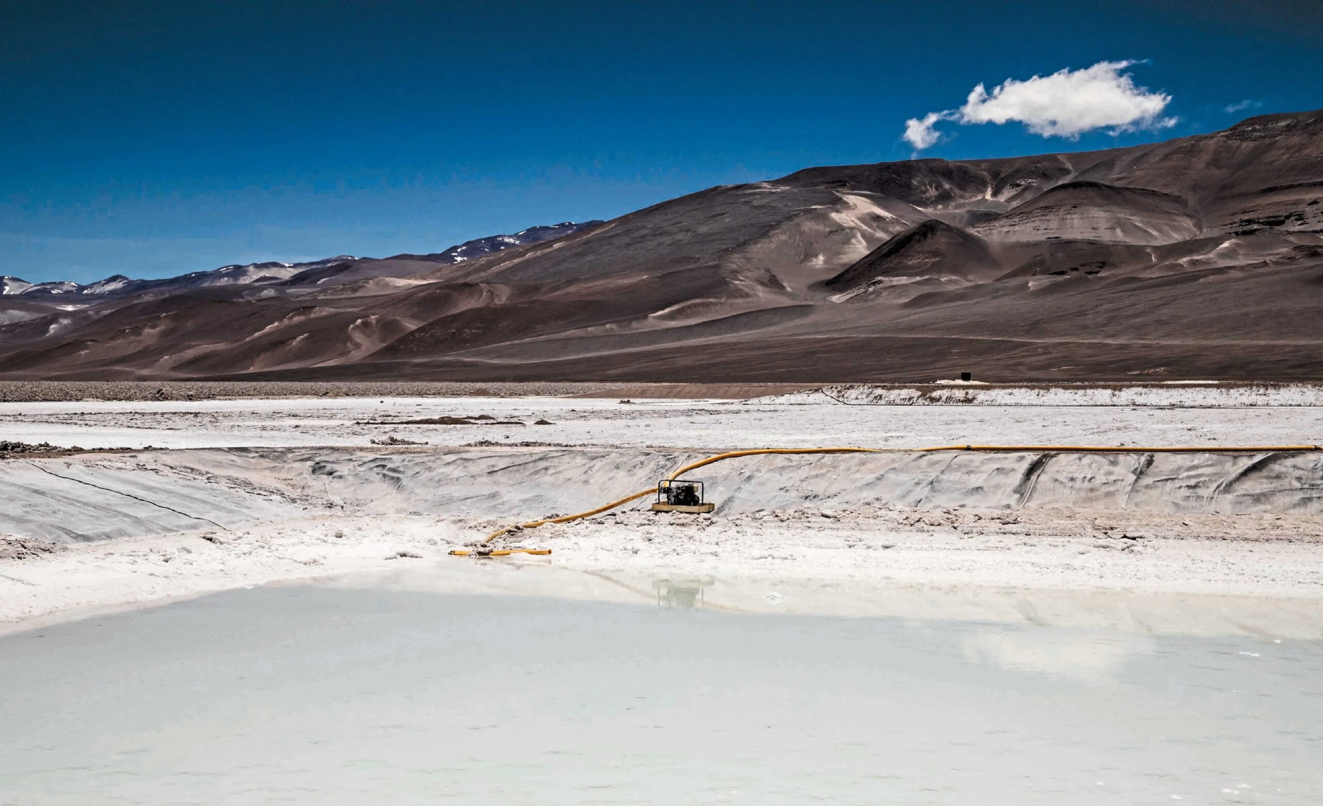 Un piccolo veicolo con tubi gialli si trova accanto a una piana di sale o a uno stagno di evaporazione pallido e riflettente, sullo sfondo di montagne aride e marroni e di un cielo blu intenso con un'unica nuvola bianca.