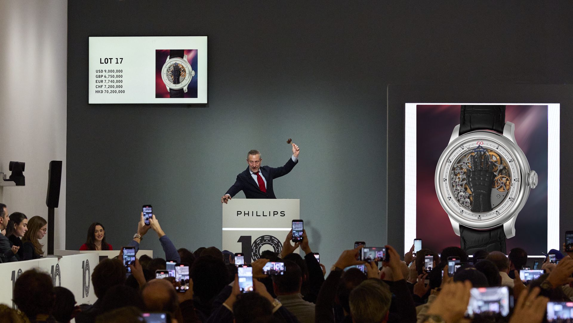 Un banditore alza il martelletto durante un'asta Phillips mentre i partecipanti scattano foto. Un grande schermo e un display mostrano un orologio da polso di lusso, con prezzi d'asta indicati in varie valute.