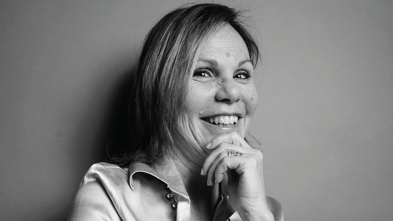 Foto in bianco e nero di una donna sorridente con i capelli lunghi fino alle spalle, che indossa una camicetta di raso e appoggia il mento sulla mano mentre guarda la macchina fotografica, riflettendo la qualità Comgest sia nello stile che nell'eleganza.