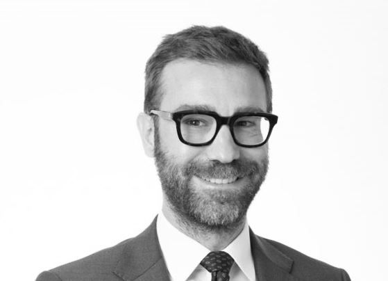 Un uomo con barba e occhiali, in giacca e cravatta, sorride su uno sfondo bianco.