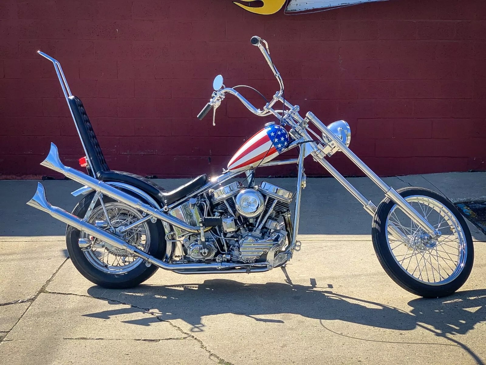Una moto chopper custom con forcella anteriore allungata, manubrio alto e serbatoio rosso, bianco e blu con stelle bianche; parcheggiata in una strada illuminata dal sole contro un muro di mattoni rossi.