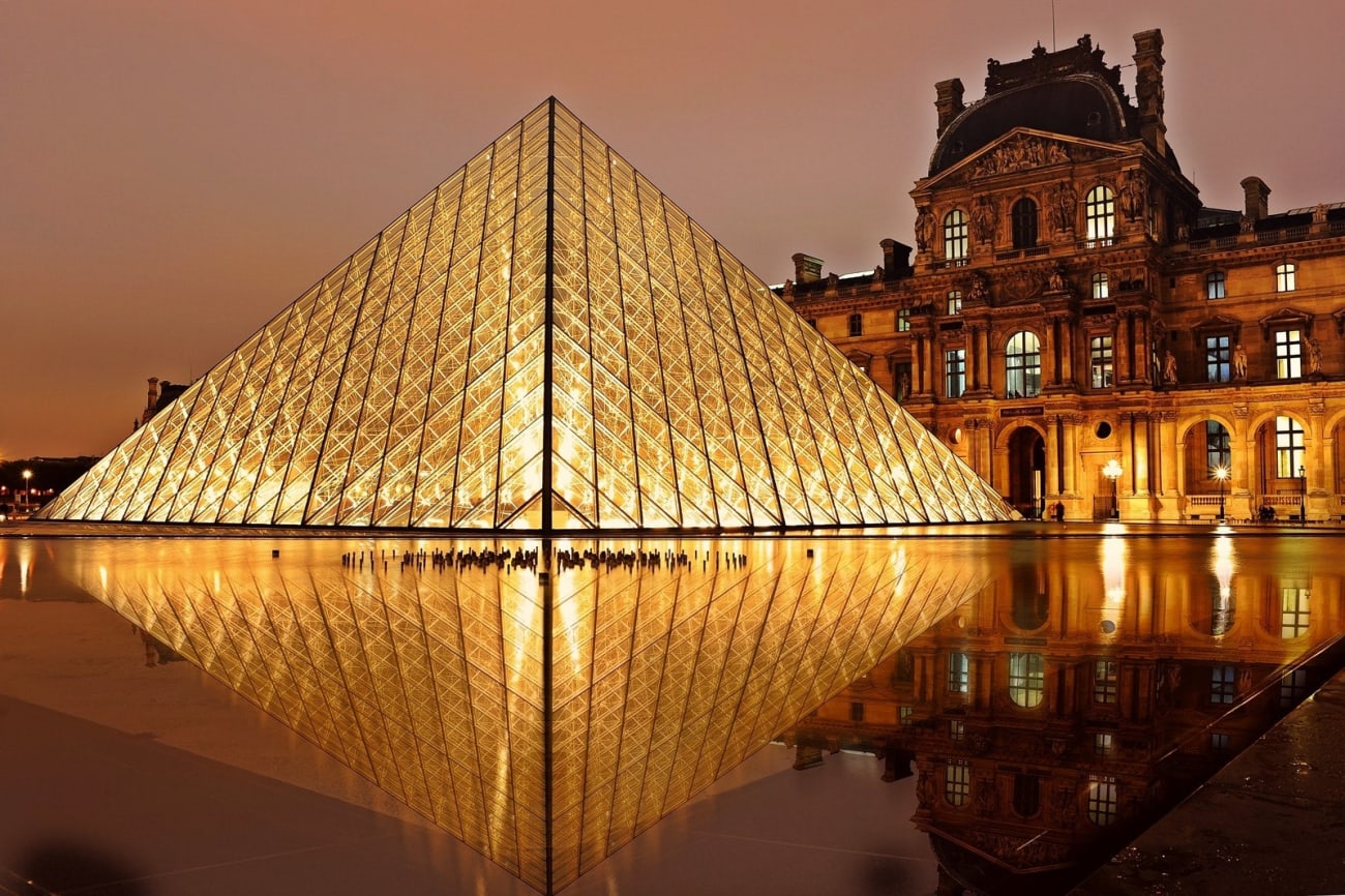 La piramide di vetro illuminata del Museo del Louvre di notte, che si riflette in uno specchio d'acqua, con l'edificio storico del museo illuminato sullo sfondo sotto un cielo arancione.
