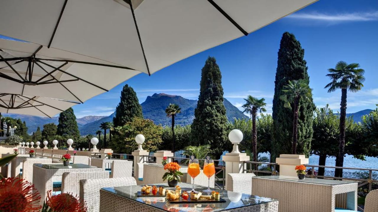 Outdoor terrace with white wicker tables and chairs, orange drinks, snacks, and flower vases; large umbrellas provide shade. Tall trees, a lake, and distant mountains are visible under a clear blue sky in the background.