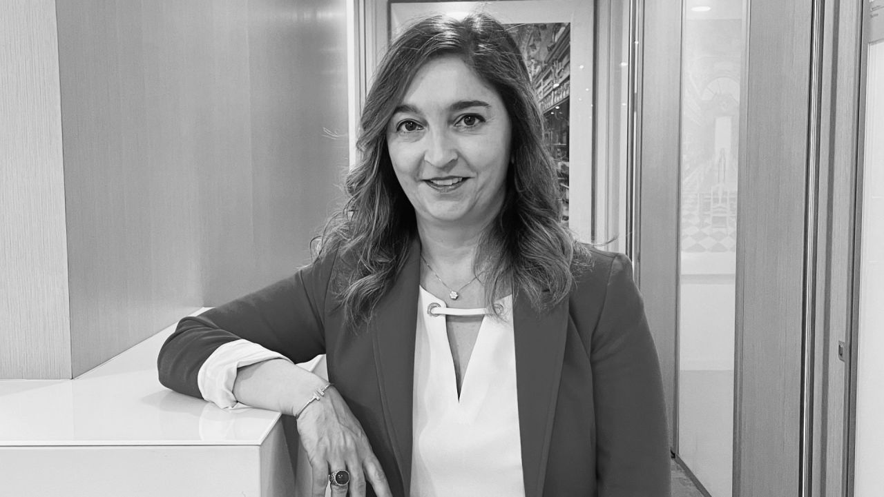 Una donna con i capelli mossi, che indossa un blazer e un top chiaro, sta in casa appoggiata a un piano di lavoro e sorride alla telecamera. L'ambiente sembra essere il corridoio di un ufficio moderno. La foto è in bianco e nero.