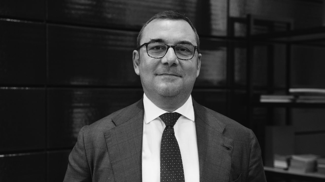 Un uomo in giacca e cravatta e con gli occhiali sorride leggermente mentre si trova in casa davanti a una parete e a scaffali di piastrelle scure. L'immagine è in bianco e nero.