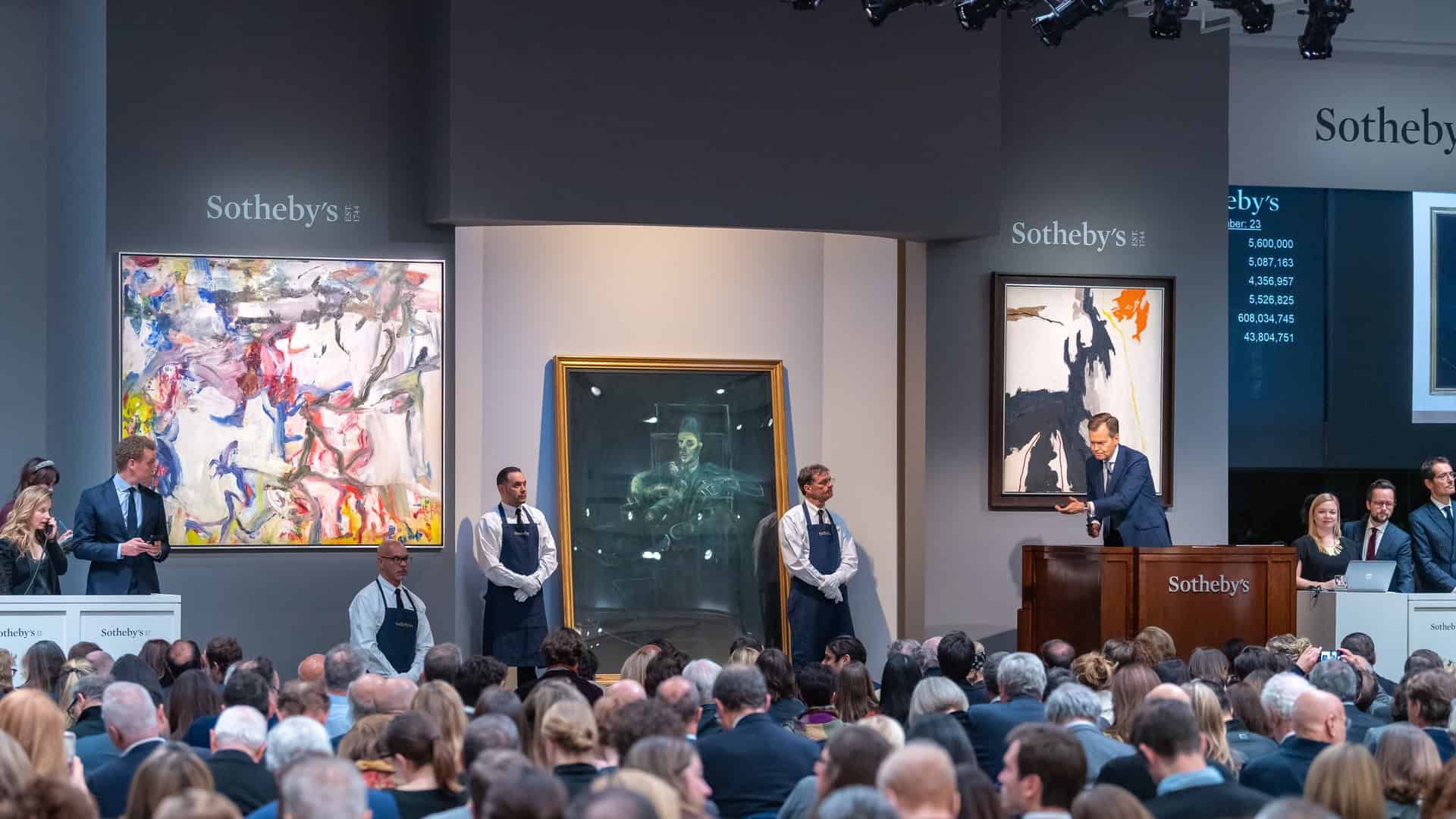Un banditore conduce un'asta d'arte di Sotheby's in una grande sala piena di partecipanti seduti. Dietro di lui ci sono due dipinti astratti e alcuni membri dello staff in piedi. Su uno schermo digitale vengono visualizzate le offerte attuali.