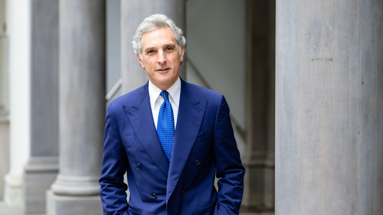 Un uomo con i capelli grigi, in doppiopetto blu e cravatta a fantasia blu, si trova di fronte a grandi colonne di pietra e guarda la telecamera con un leggero sorriso.