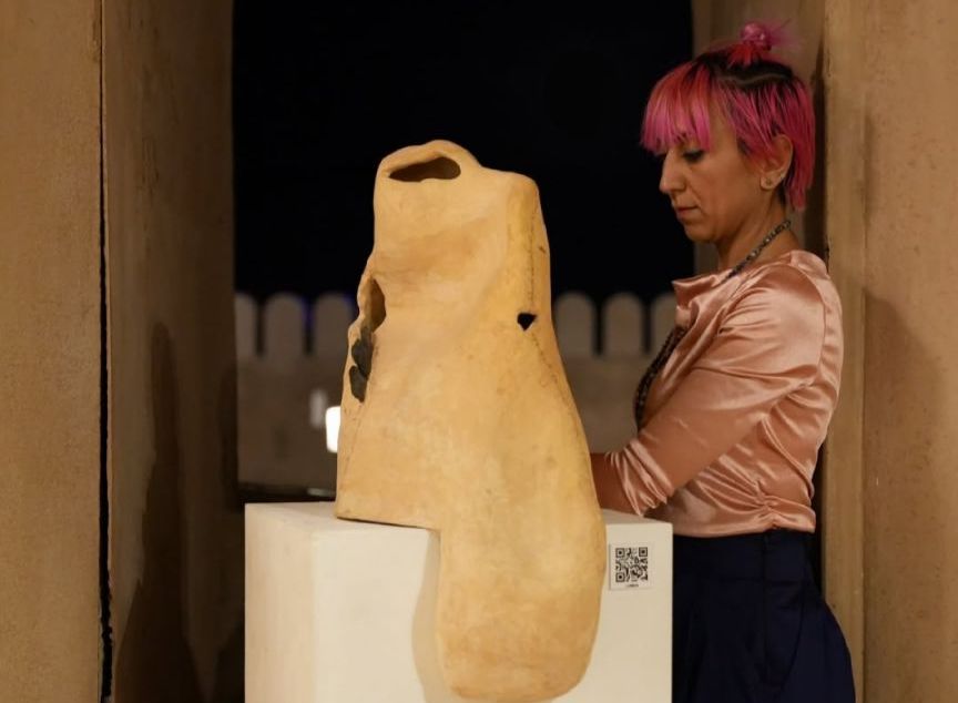 Una donna con i capelli corti rosa e una camicetta di raso rosa si trova accanto a una scultura astratta beige esposta su un piedistallo bianco, con il muro di una fortezza visibile sullo sfondo di notte.