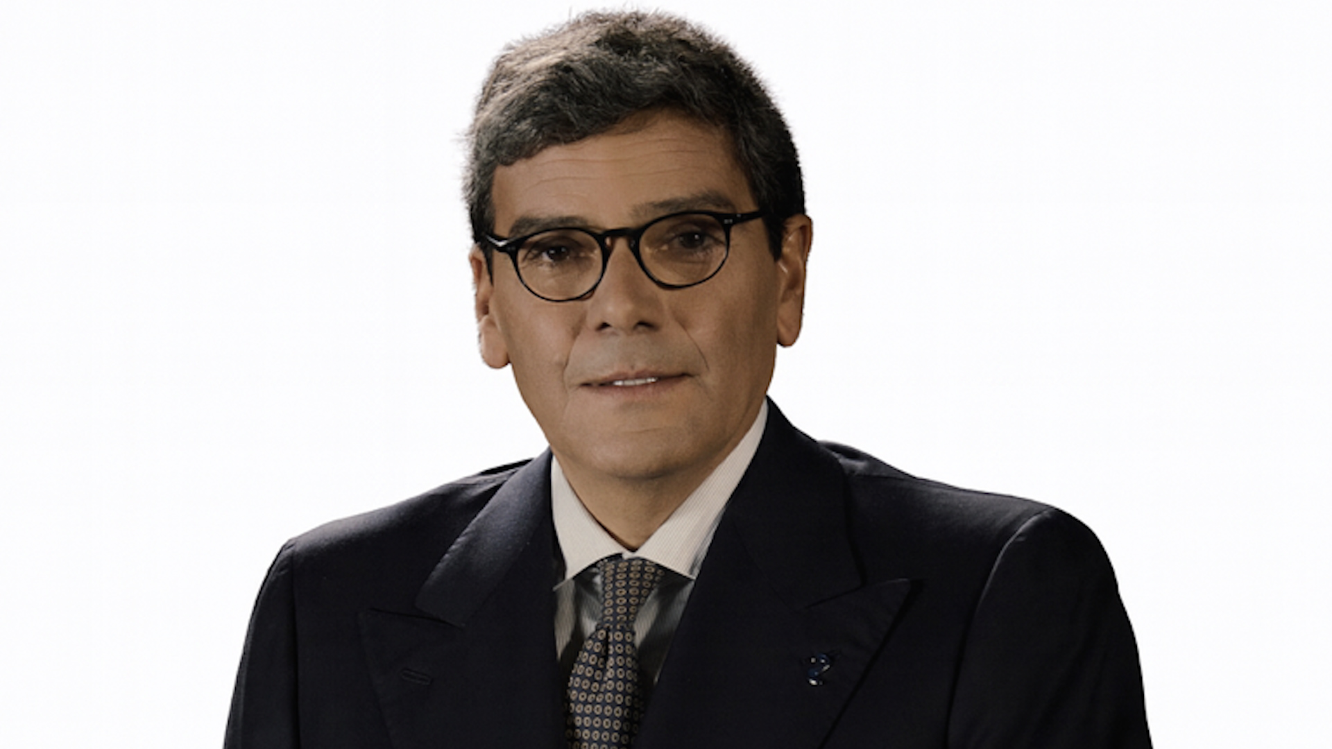Nicola Riello con capelli corti e occhiali, vestito con abito scuro, camicia bianca e cravatta a fantasia, posa su un semplice sfondo bianco. Guarda direttamente la telecamera con un'espressione neutra, rappresentando la professionalità di Private Equity Italia.