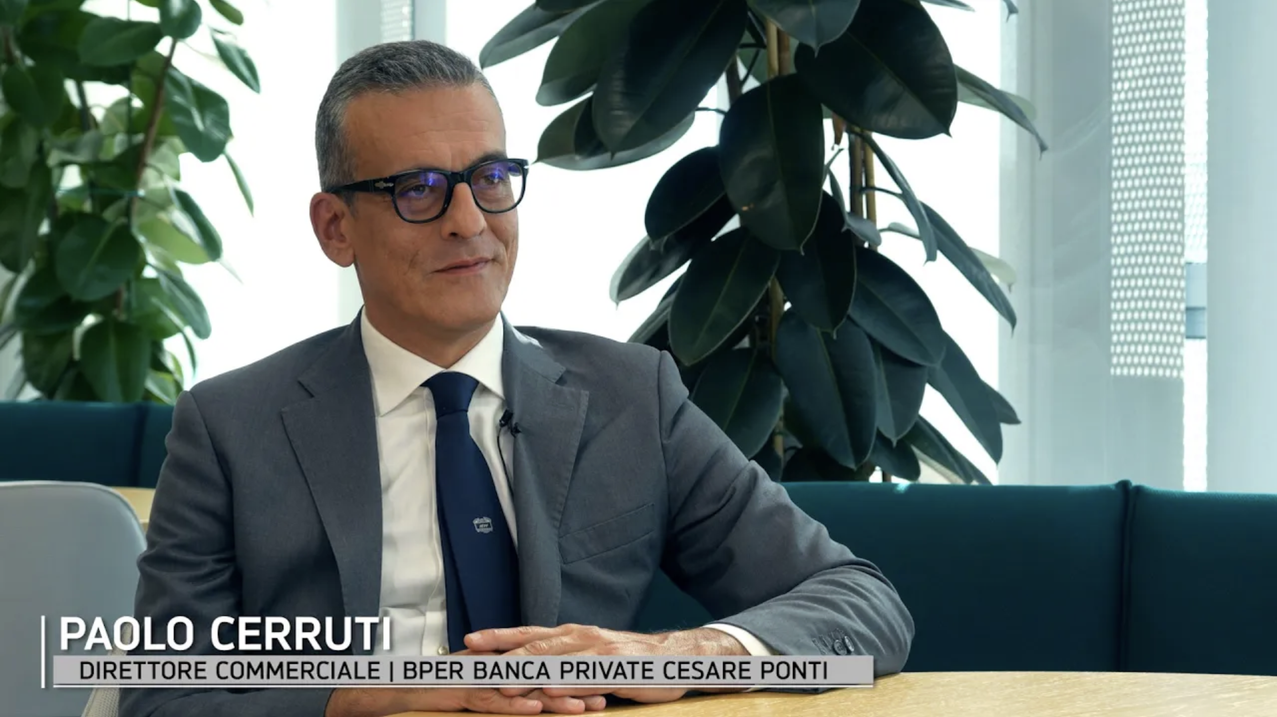 Un uomo in abito grigio, camicia bianca e cravatta blu siede a un tavolo in un ufficio moderno con piante e grandi finestre. Il testo dell'immagine recita: "Paolo Cerruti, Direttore Commerciale | BPER Banca Private Cesare Ponti".