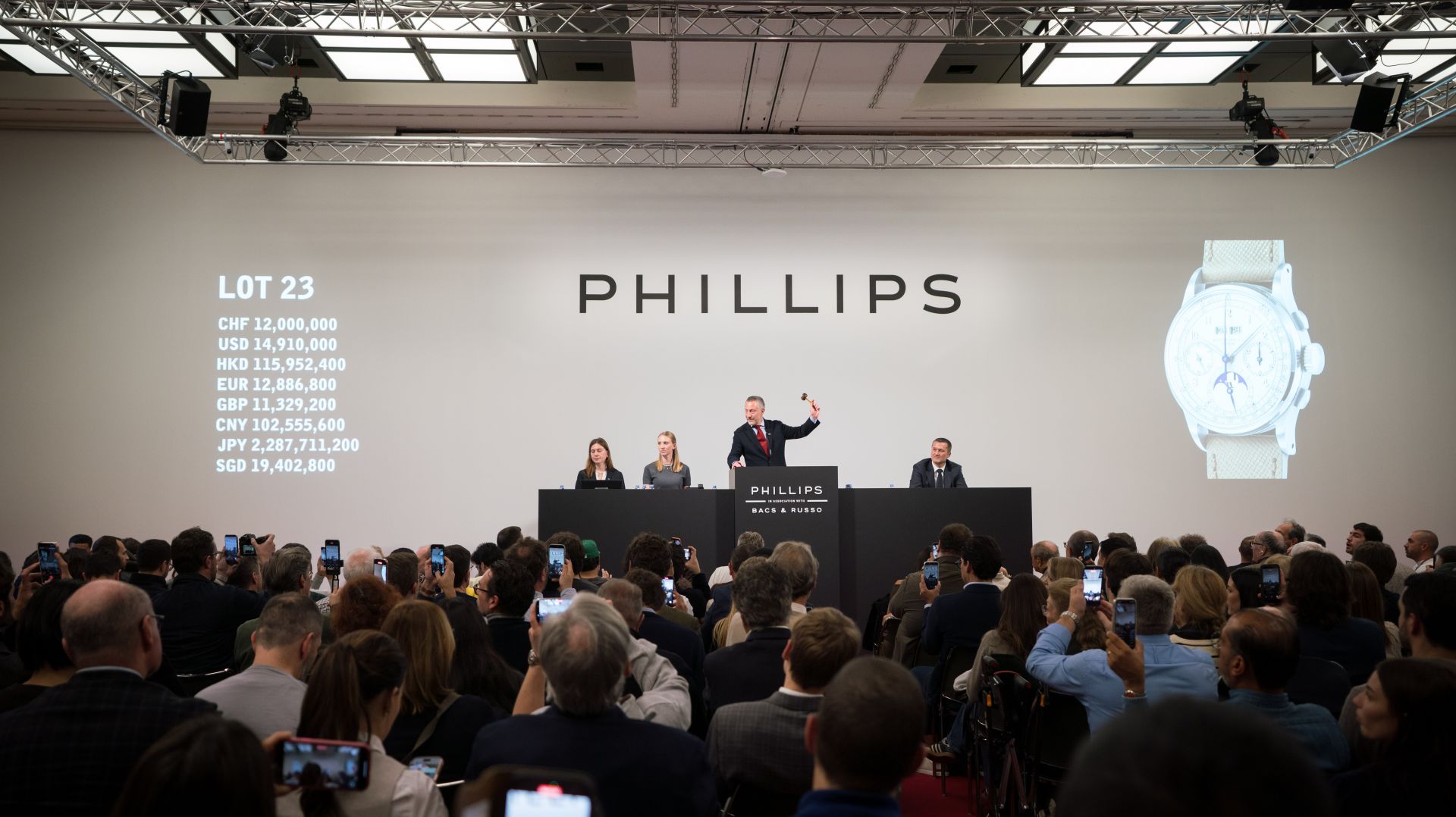 Una sala d'asta affollata presso Phillips Watches. Un banditore fa dei gesti mentre tre membri dello staff siedono su un podio. Su uno schermo vengono visualizzati il lotto 23 e le offerte attuali in varie valute. Molti partecipanti stanno scattando foto.