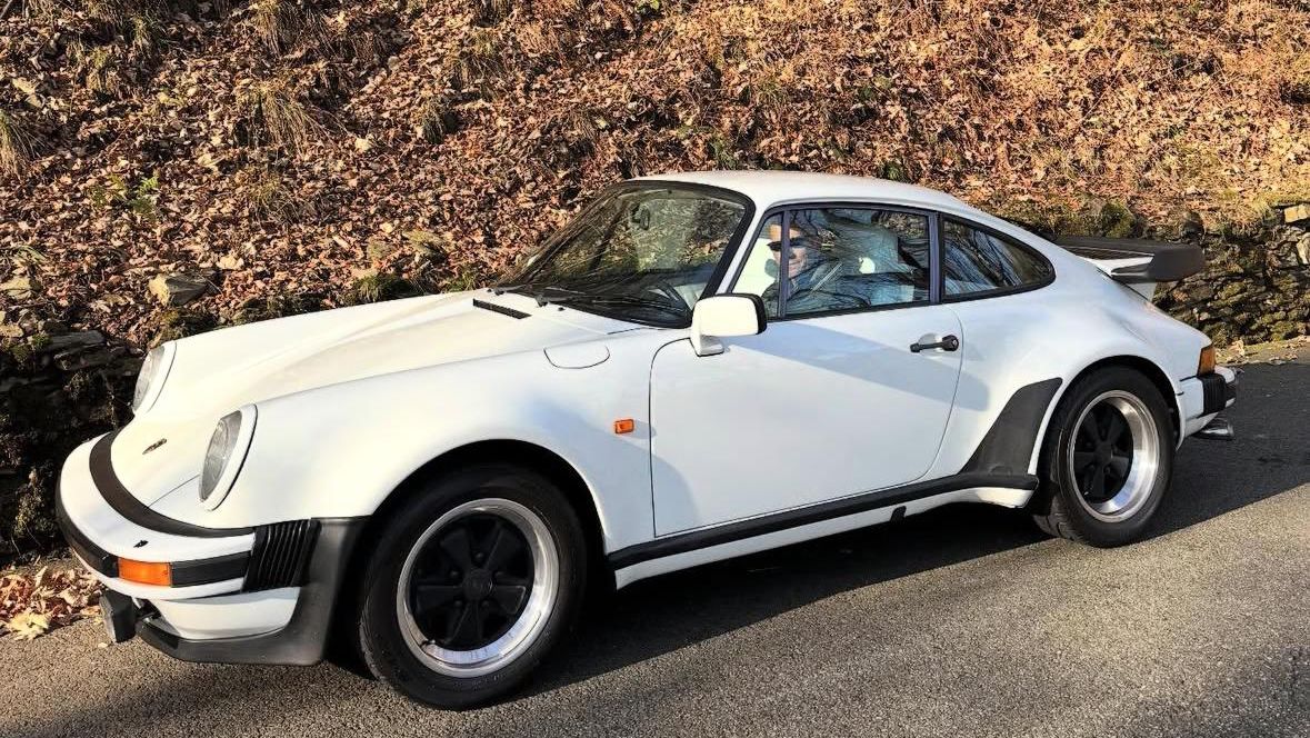 A classic white Porsche 911 Turbo parked on a road beside a stone wall and autumn leaves, with a person seated in the driver’s seat.