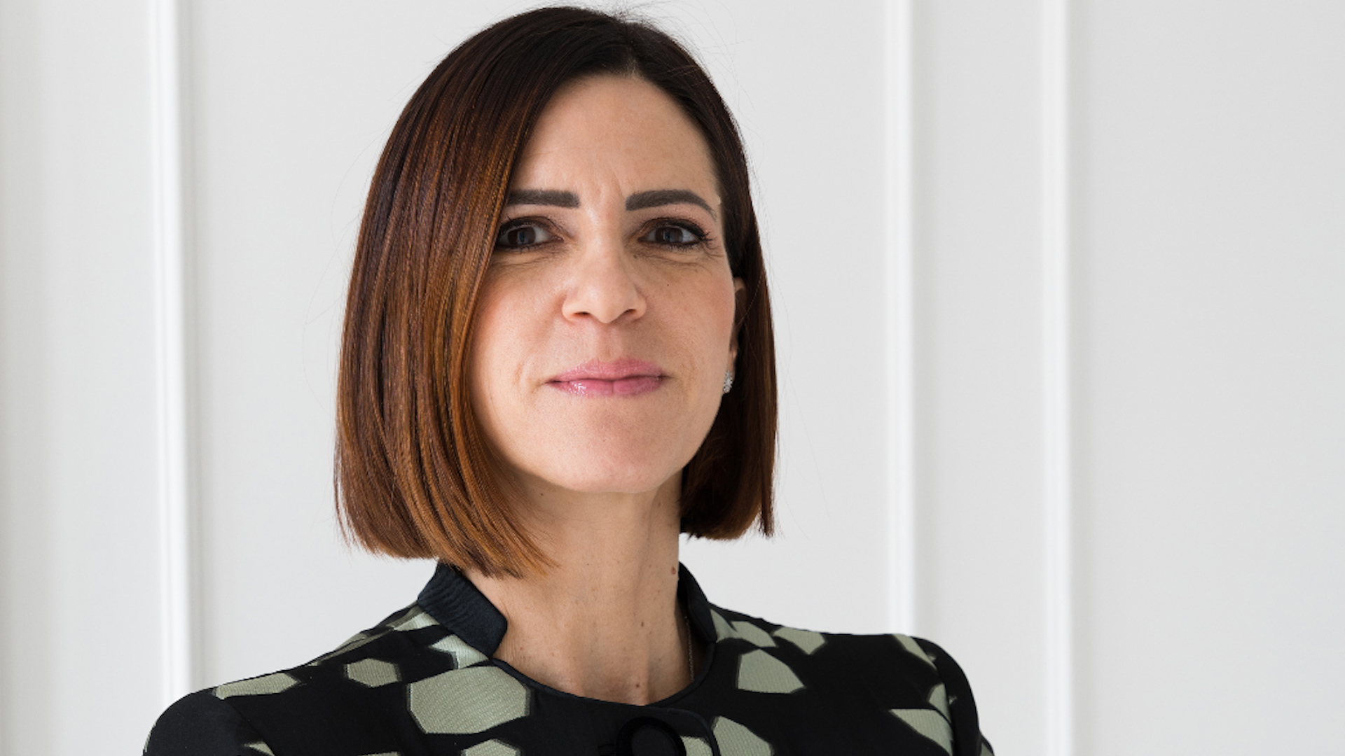 Roberta Vinciguerra con capelli castani lisci e lunghi fino alle spalle e una giacca nera a fantasia si trova di fronte a una parete bianca e guarda con sicurezza verso la telecamera, cogliendo lo spirito professionale che spesso si respira nell'ufficio di famiglia Vedalia.