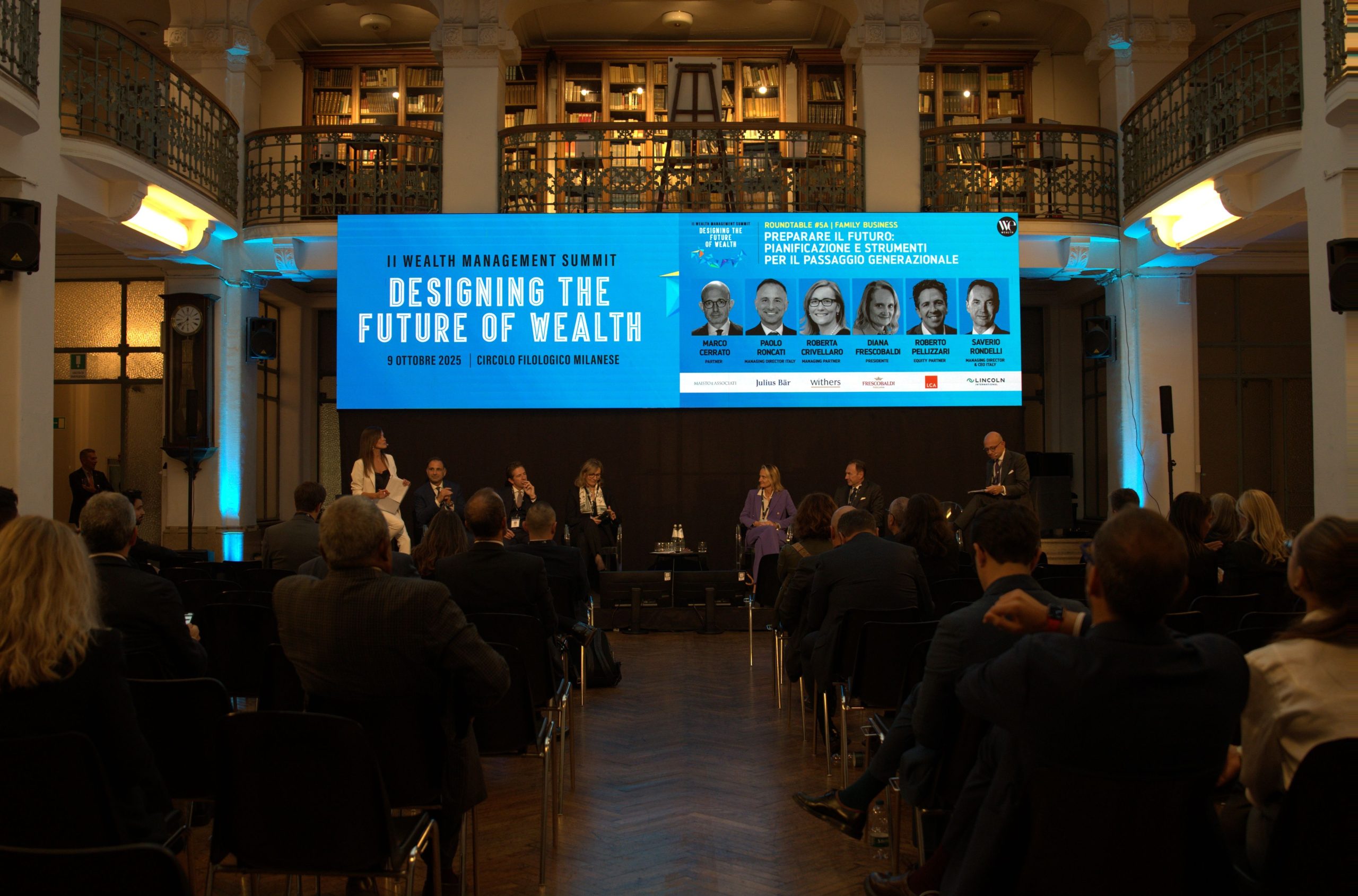 Una sala conferenze con partecipanti seduti ascolta un gruppo di relatori sul palco. Alle loro spalle, un grande schermo digitale mostra i dettagli dell'evento: "Designing the Future of Wealth and Passaggio Generazionale", con nomi e foto dei relatori.