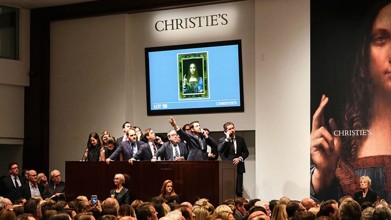 Si sta svolgendo un'asta da Christie's, con diversi banditori e personale sul podio. Una grande folla sta guardando mentre il "Salvator Mundi", un tempo legato a Bouvier, viene visualizzato su uno schermo alle loro spalle.