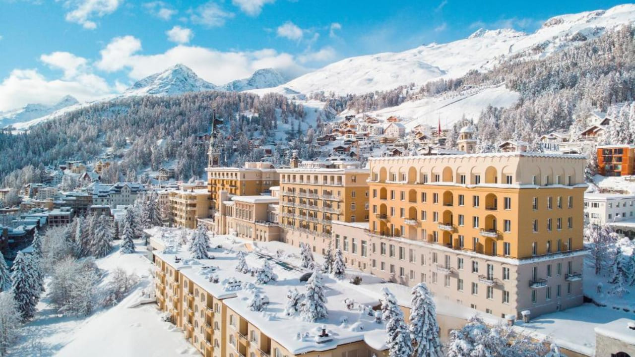 A large, elegant hotel complex sits surrounded by snow-covered trees and buildings, set against a backdrop of snowy mountains and a clear blue sky.