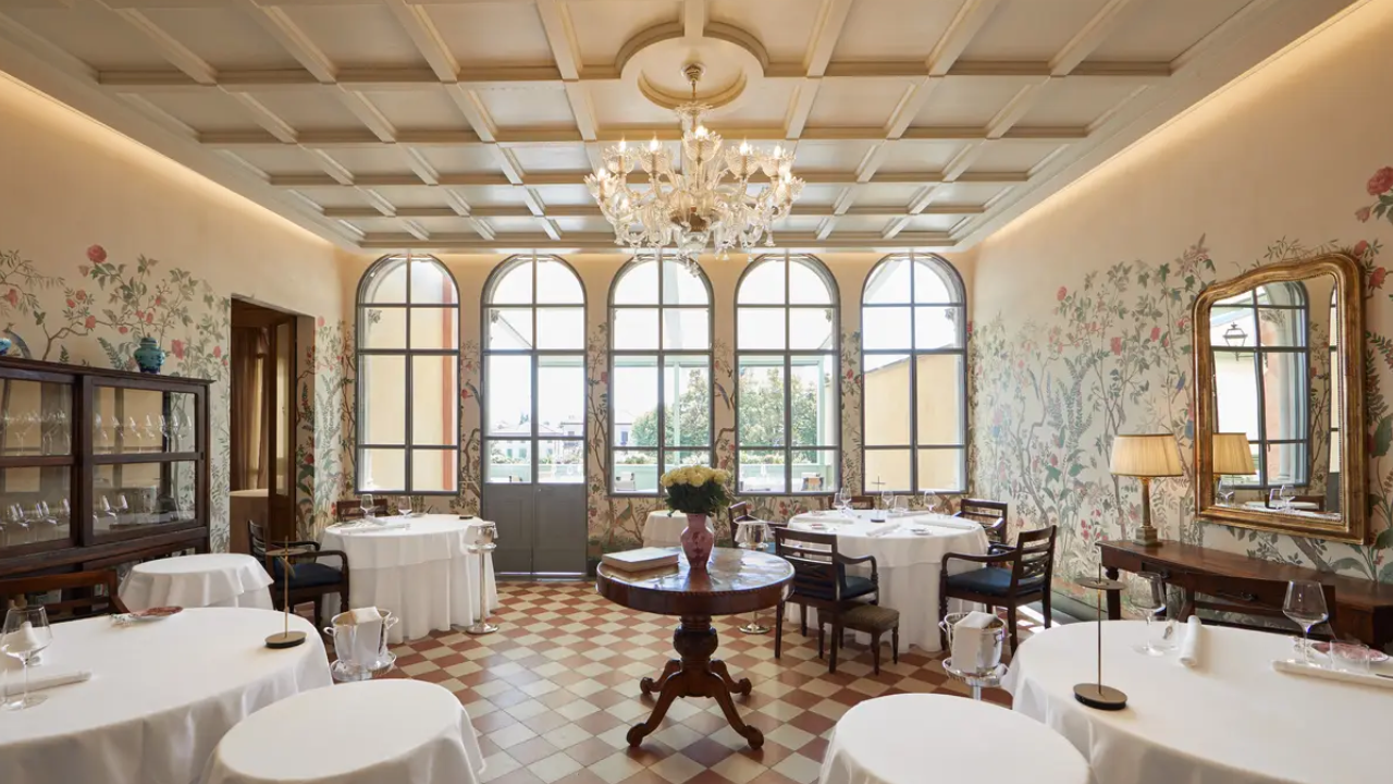 Elegante sala da pranzo del ristorante con tavoli rotondi coperti da tovaglie bianche, grandi finestre ad arco, un lampadario centrale, carta da parati floreale e mobili classici in legno su un pavimento di piastrelle a scacchi ispirato al fascino senza tempo di Trapani.