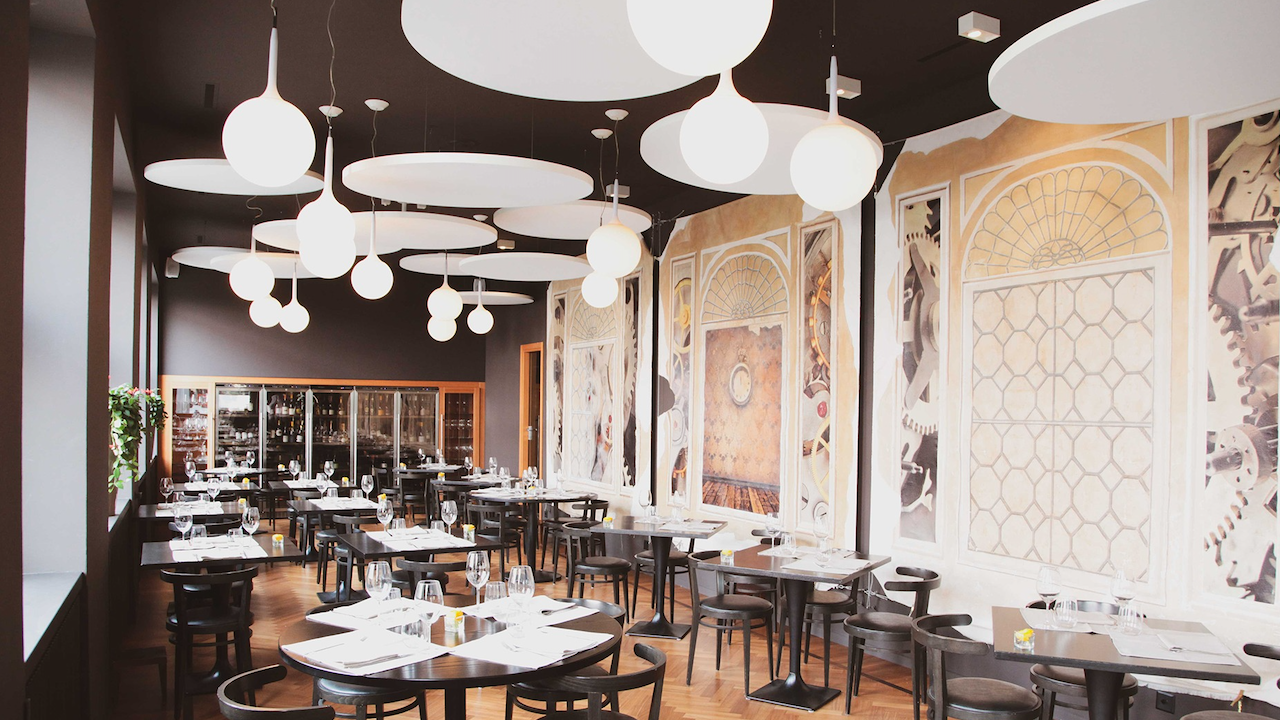 Modern restaurant interior with black tables and chairs, elegant lighting fixtures hanging from the ceiling, and large artistic murals covering one wall. Tables are set with glassware and cutlery, and the space feels bright and stylish.