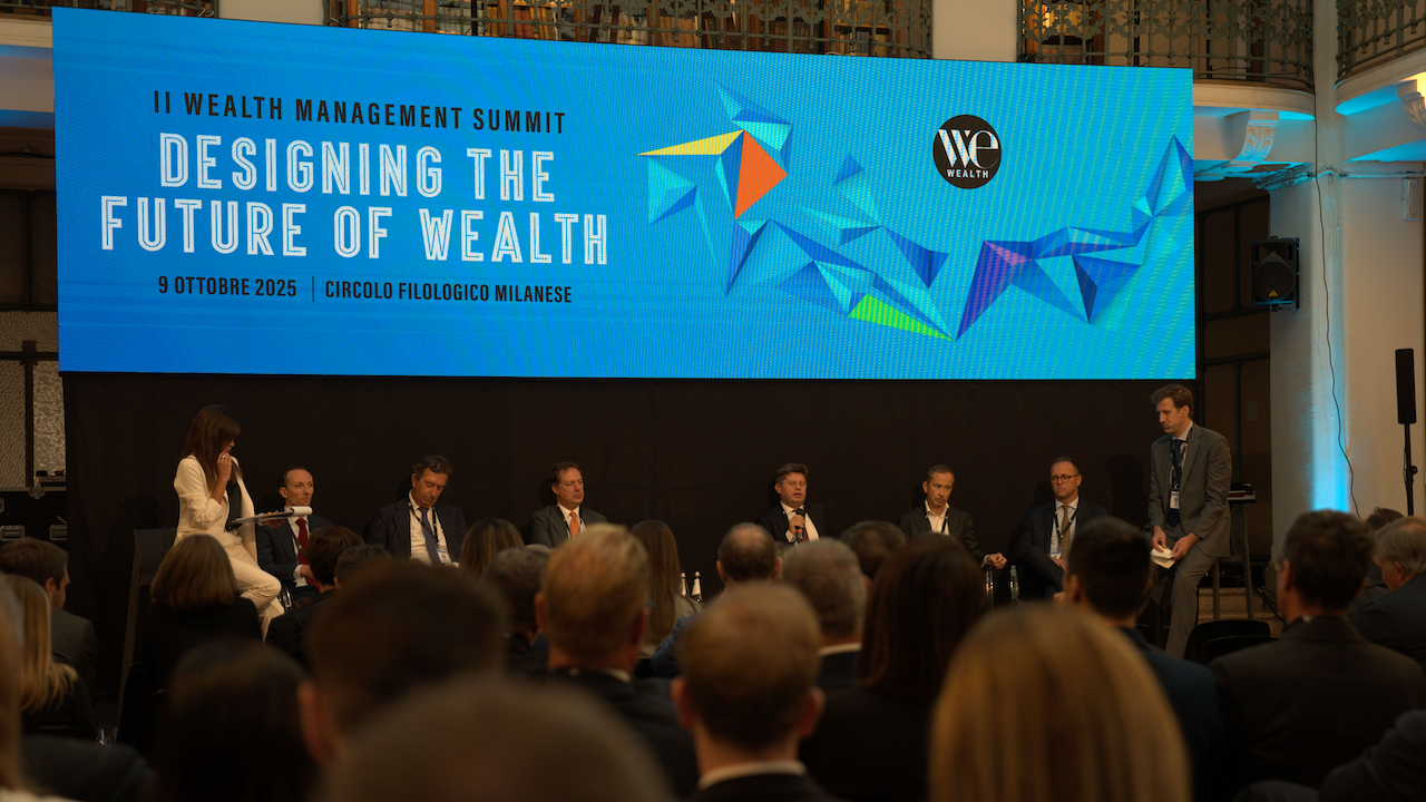 Un gruppo di relatori siede sul palco del II Wealth Management Summit, con un grande schermo blu alle loro spalle che mostra "Designing the Future of Wealth" e i dettagli dell'evento; un pubblico ascolta con attenzione.