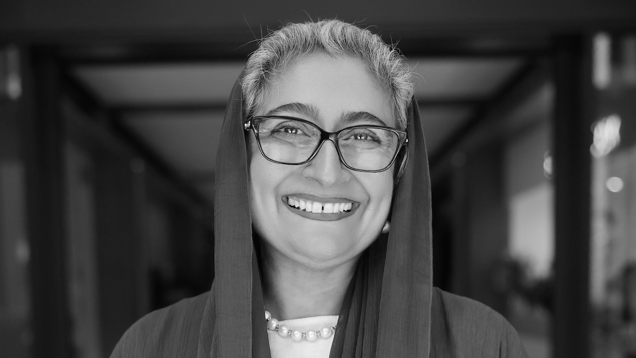 Una donna sorridente con capelli corti e grigi indossa occhiali, un foulard e una collana di perle. La foto è in bianco e nero, con uno sfondo sfocato.